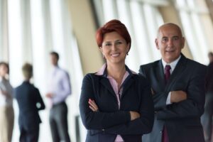 A red haired woman with a big smile wearing a suit standing in front of a well-dressed mature man